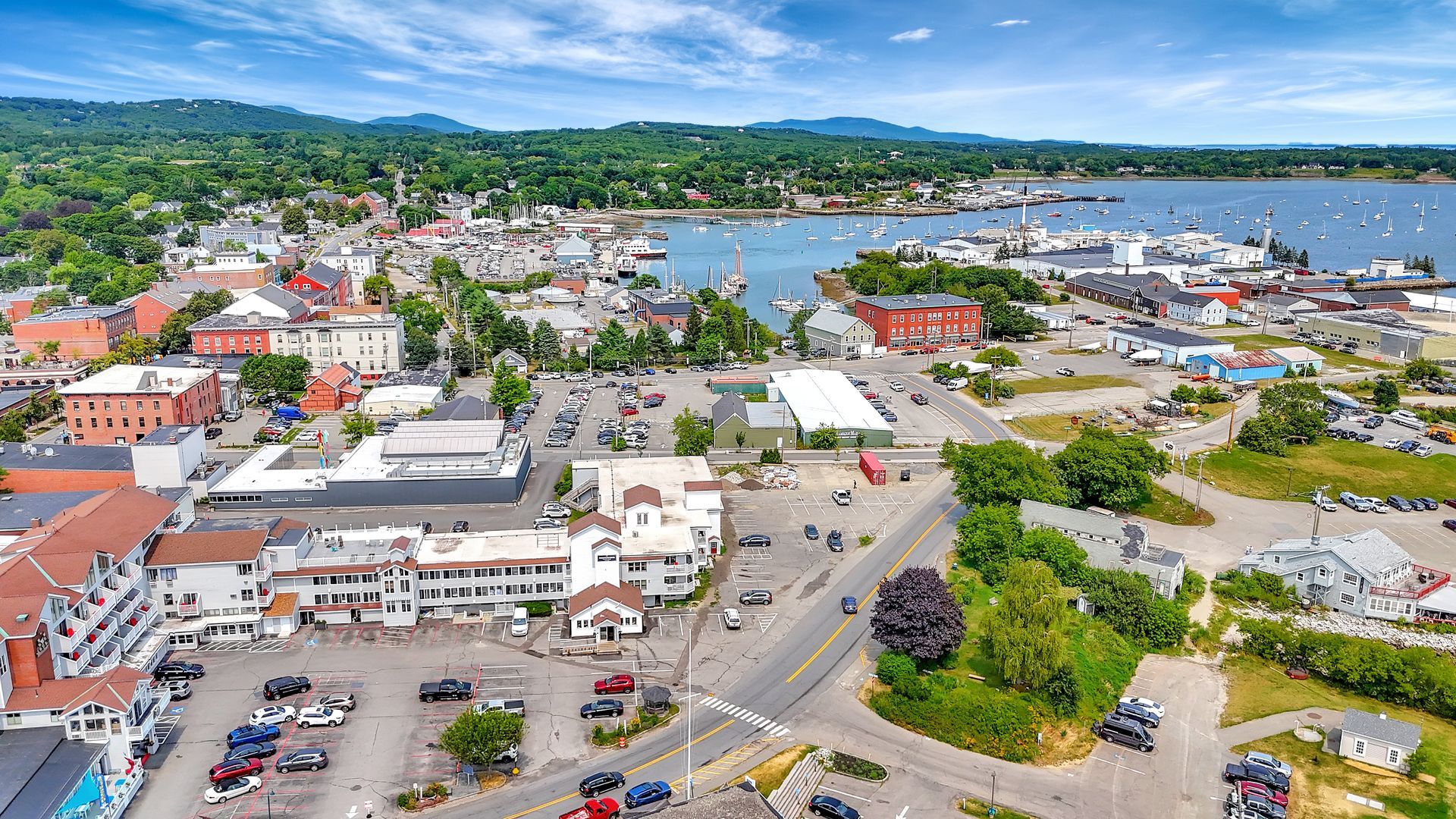 Aerial view of a coastal town with buildings, boats, and harbor, trees in the background.