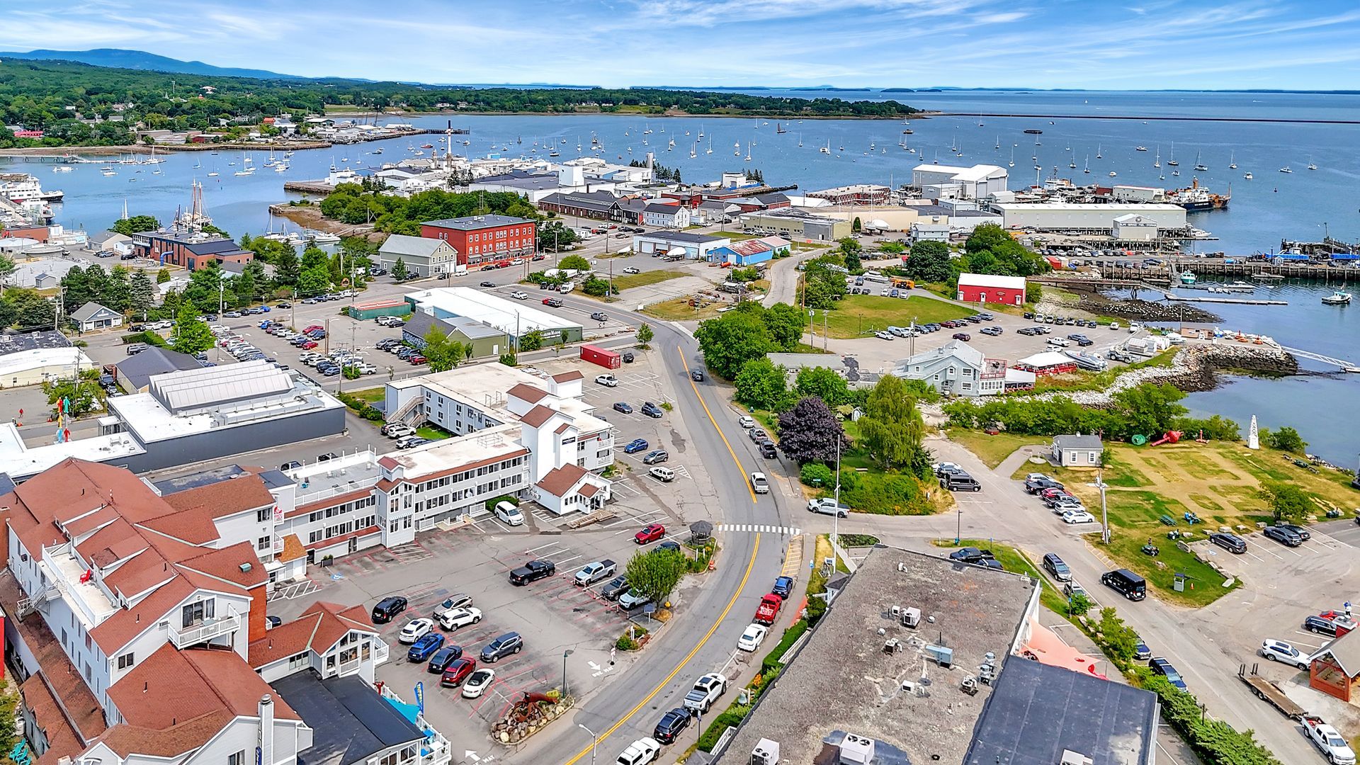 Aerial view of a coastal town with buildings, a road, docks, and boats under a blue sky.