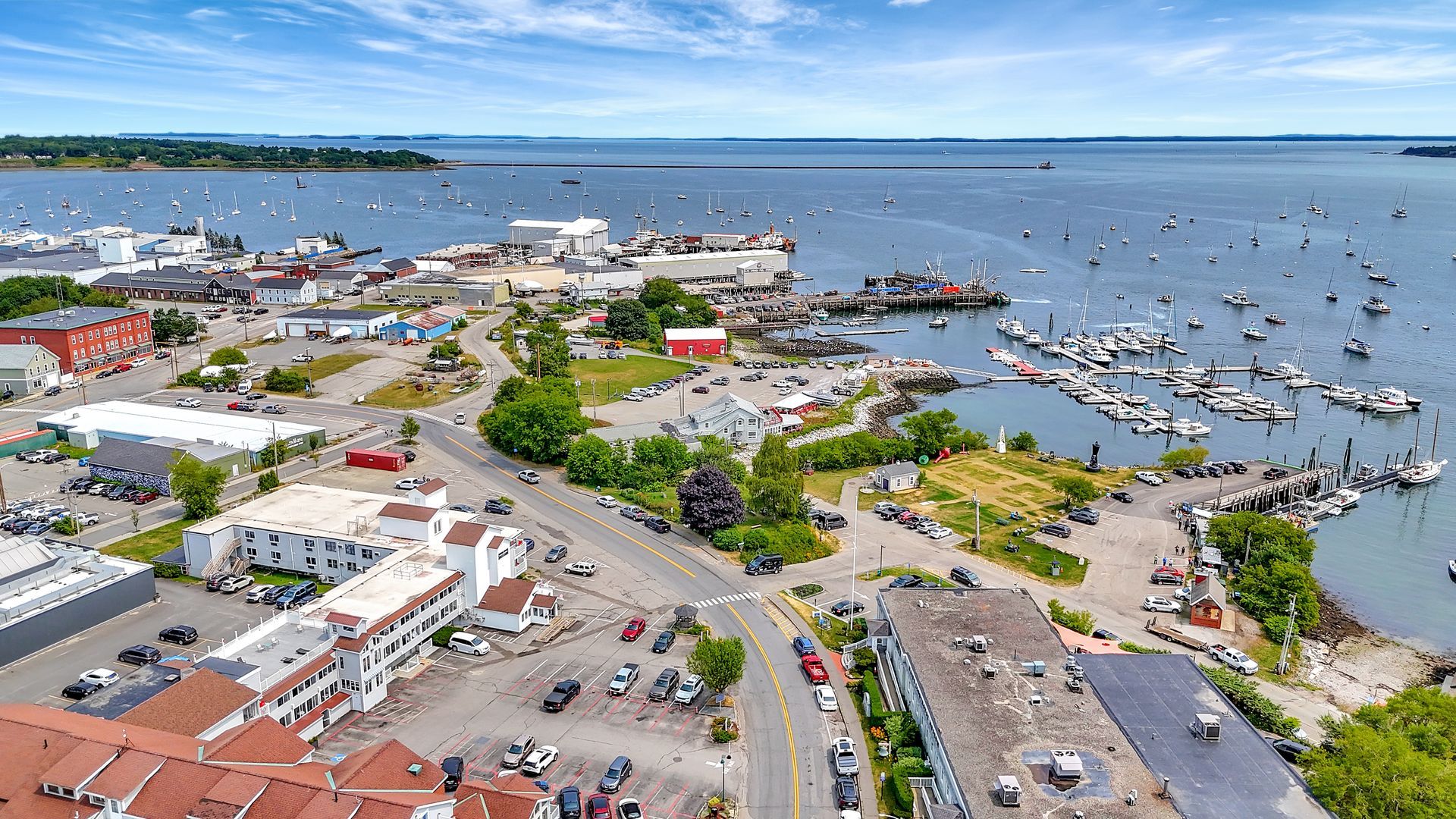 Aerial view of a coastal town with boats docked in the harbor. Buildings and roads are visible. Blue water and sky.