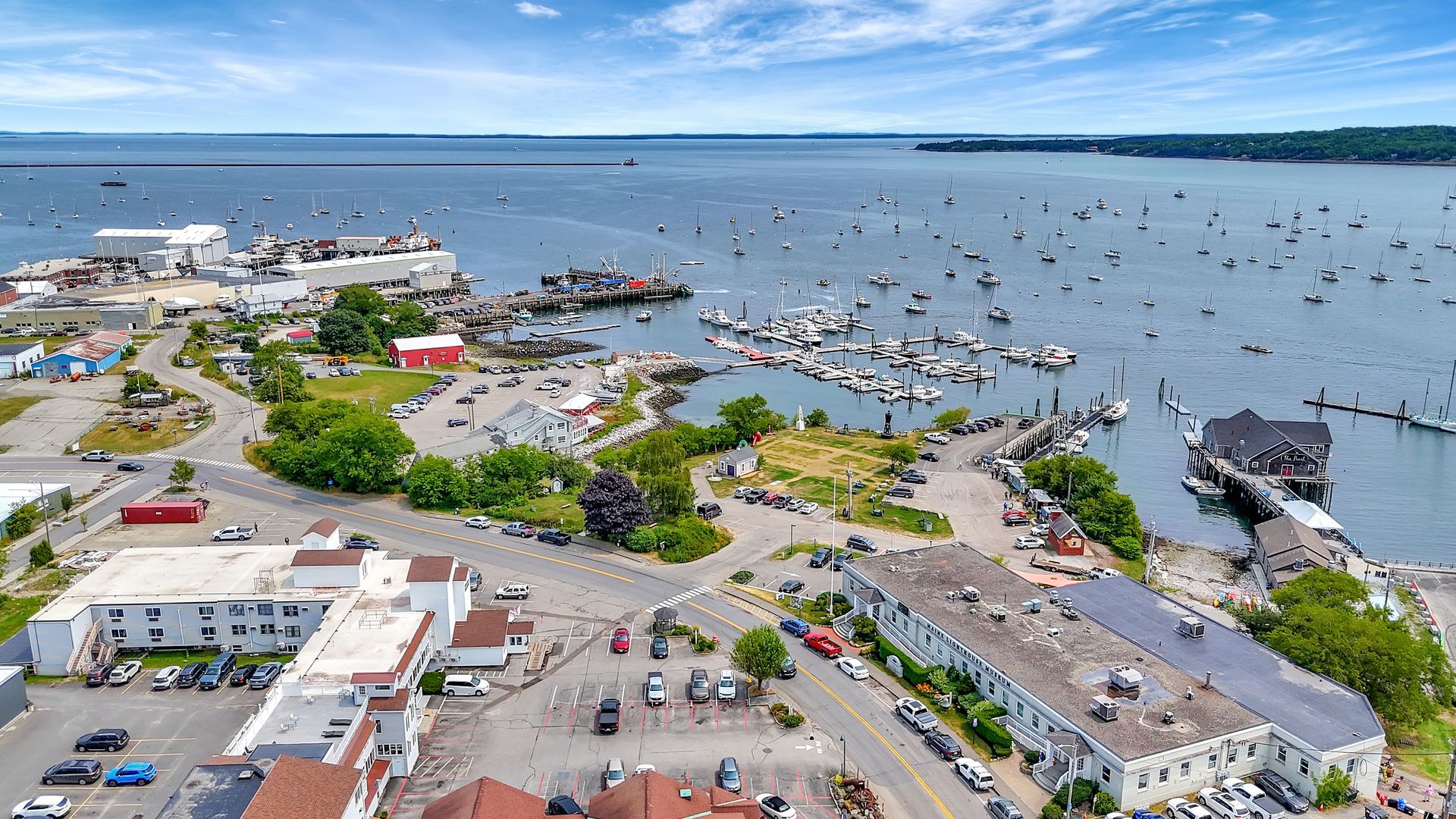 Aerial view of harbor town, boats in water, buildings along the shore under blue sky.