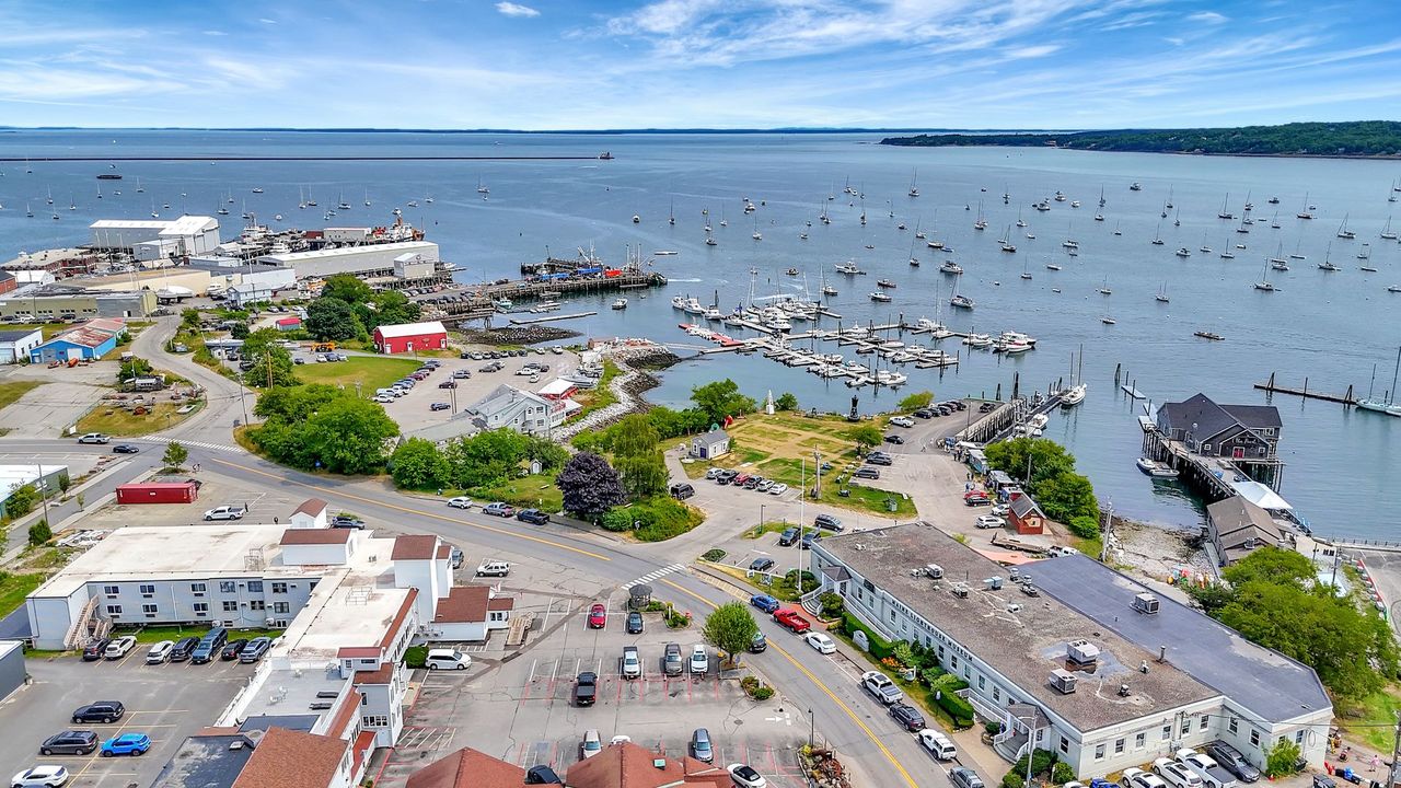 Aerial view of a harbor town with boats, buildings, and blue water under a cloudy sky.