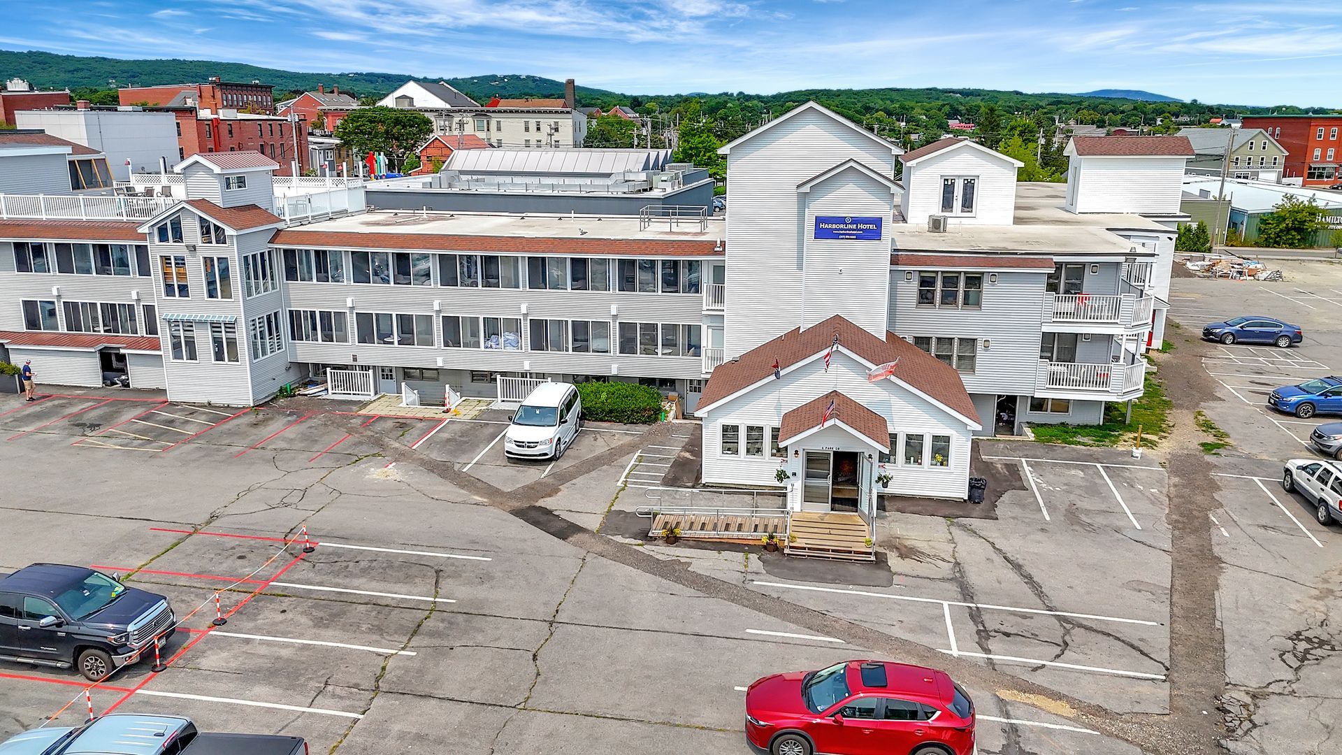 An aerial view of a gray hotel building with a parking lot filled with cars on a sunny day.