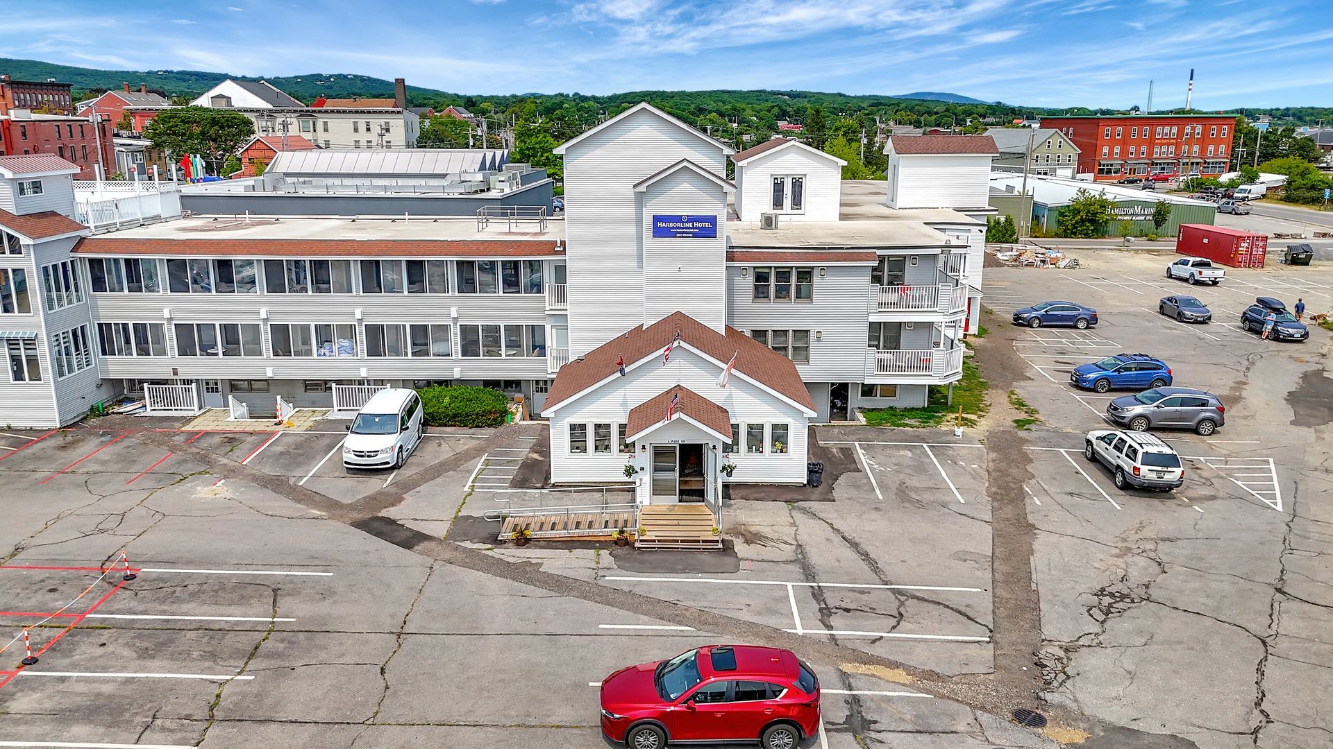 Hotel building with a parking lot and cars. Red car in the foreground. Sunny day, buildings in background.