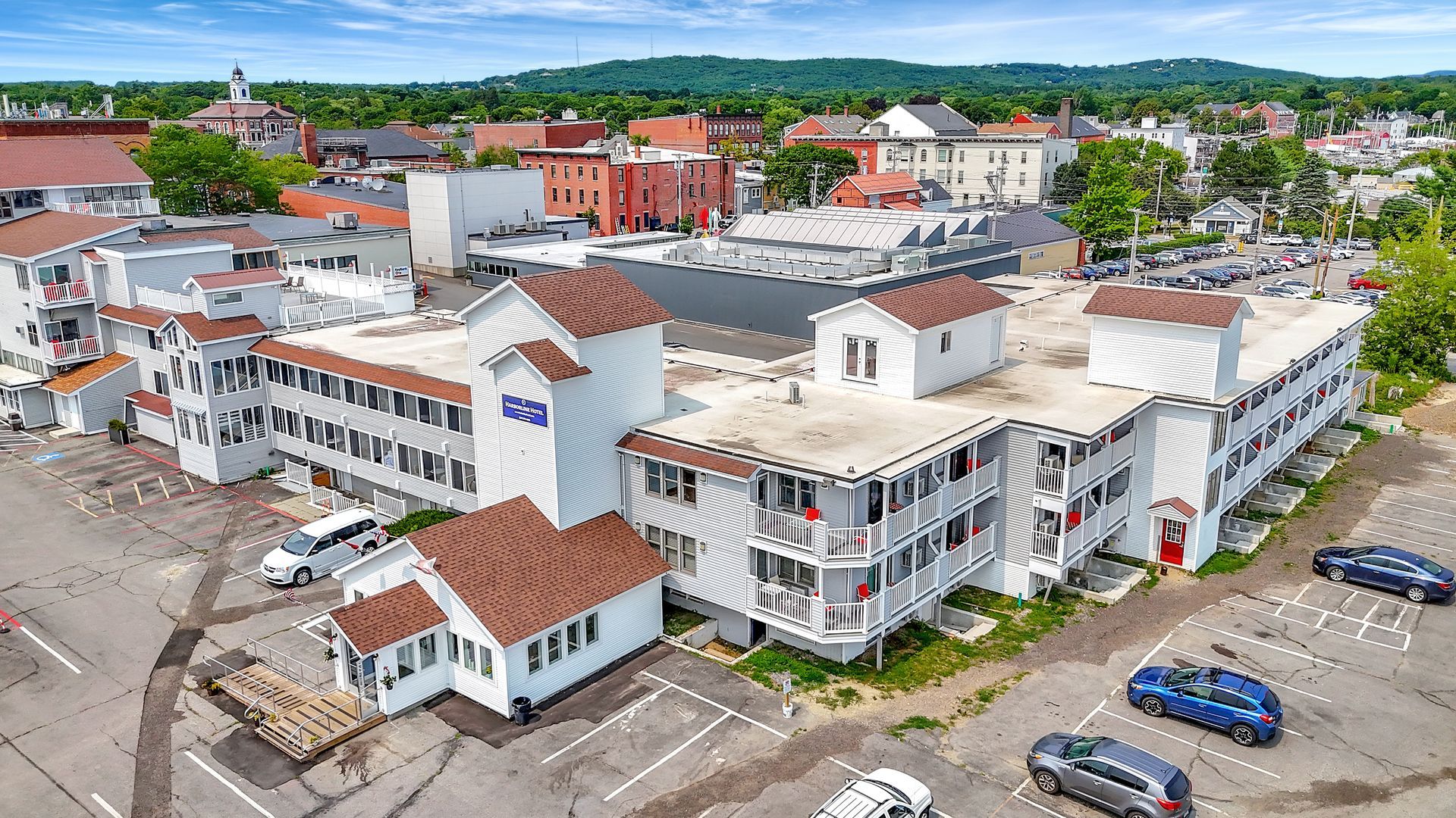 Aerial view of a white hotel with brown roofs and parking in a town with buildings and green hills in the background.