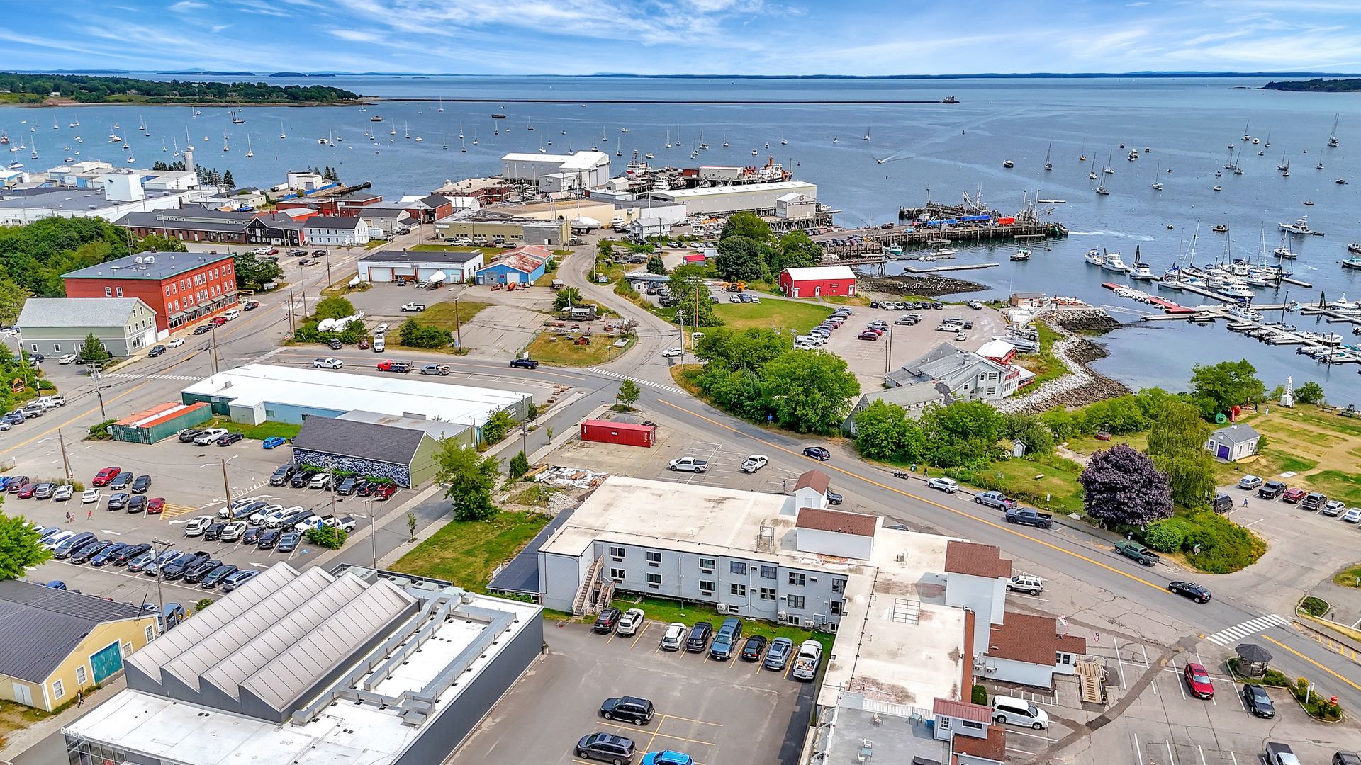 Aerial view of a harbor town with buildings, cars, boats, and a vast ocean on a sunny day.