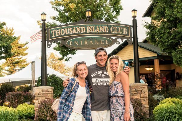 Three people are posing for a picture in front of a sign that says thousand island club entrance.