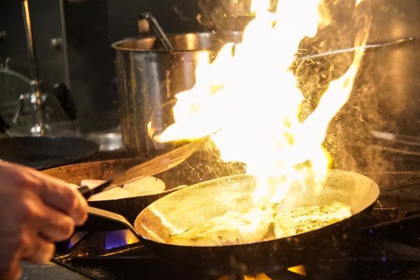 A person is cooking food in a frying pan on a stove with flames coming out of it.