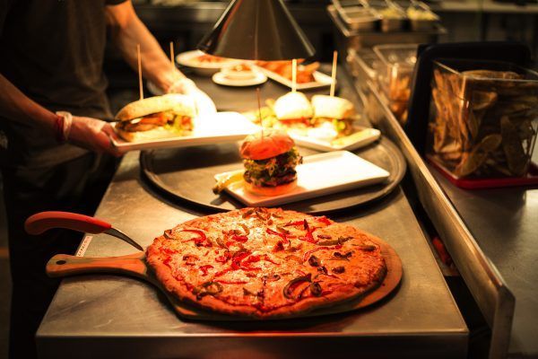 A pizza on a wooden cutting board next to a tray of hamburgers