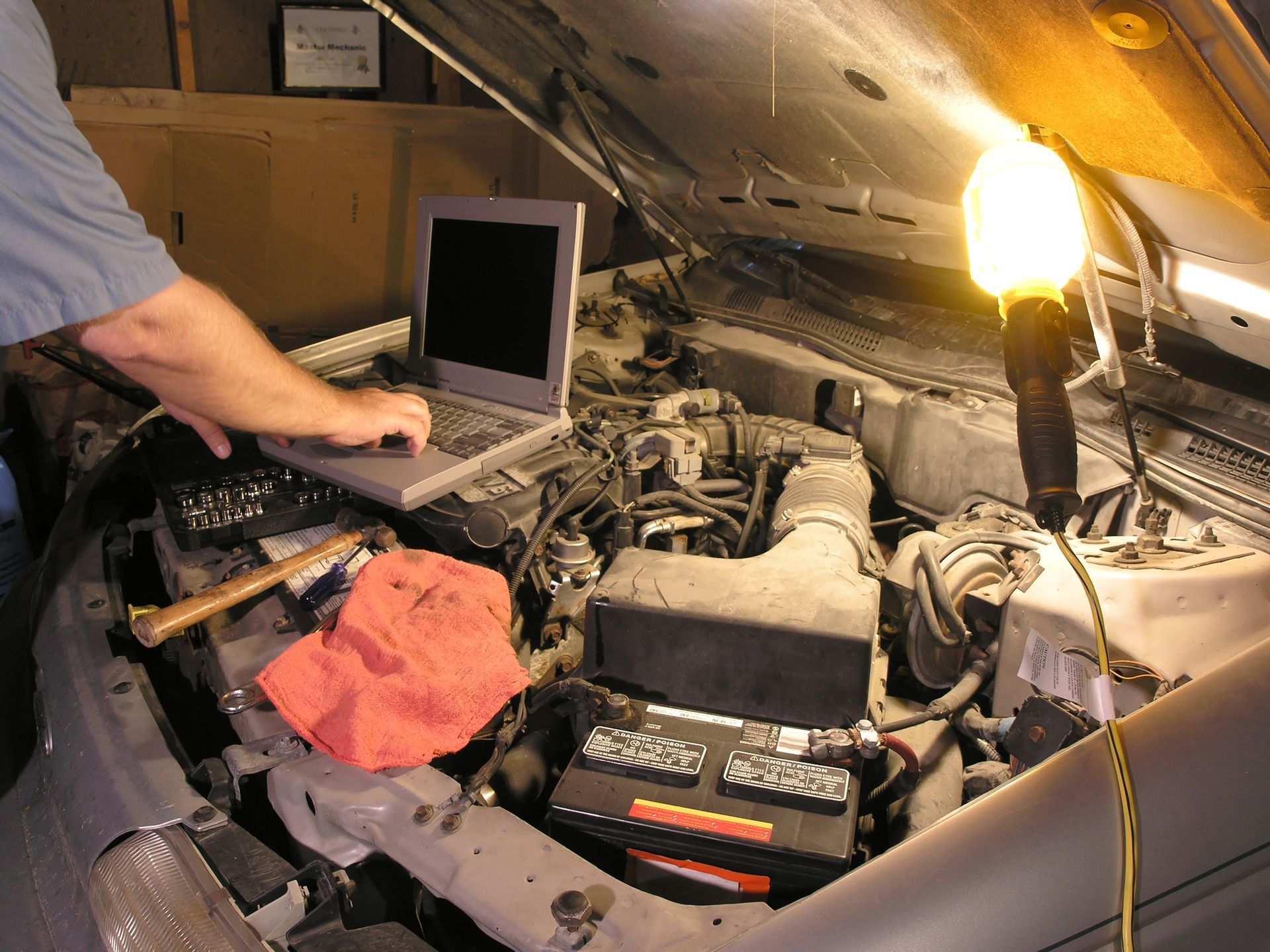 Person using laptop, possibly diagnosing a car engine. Work light illuminating the engine compartment.