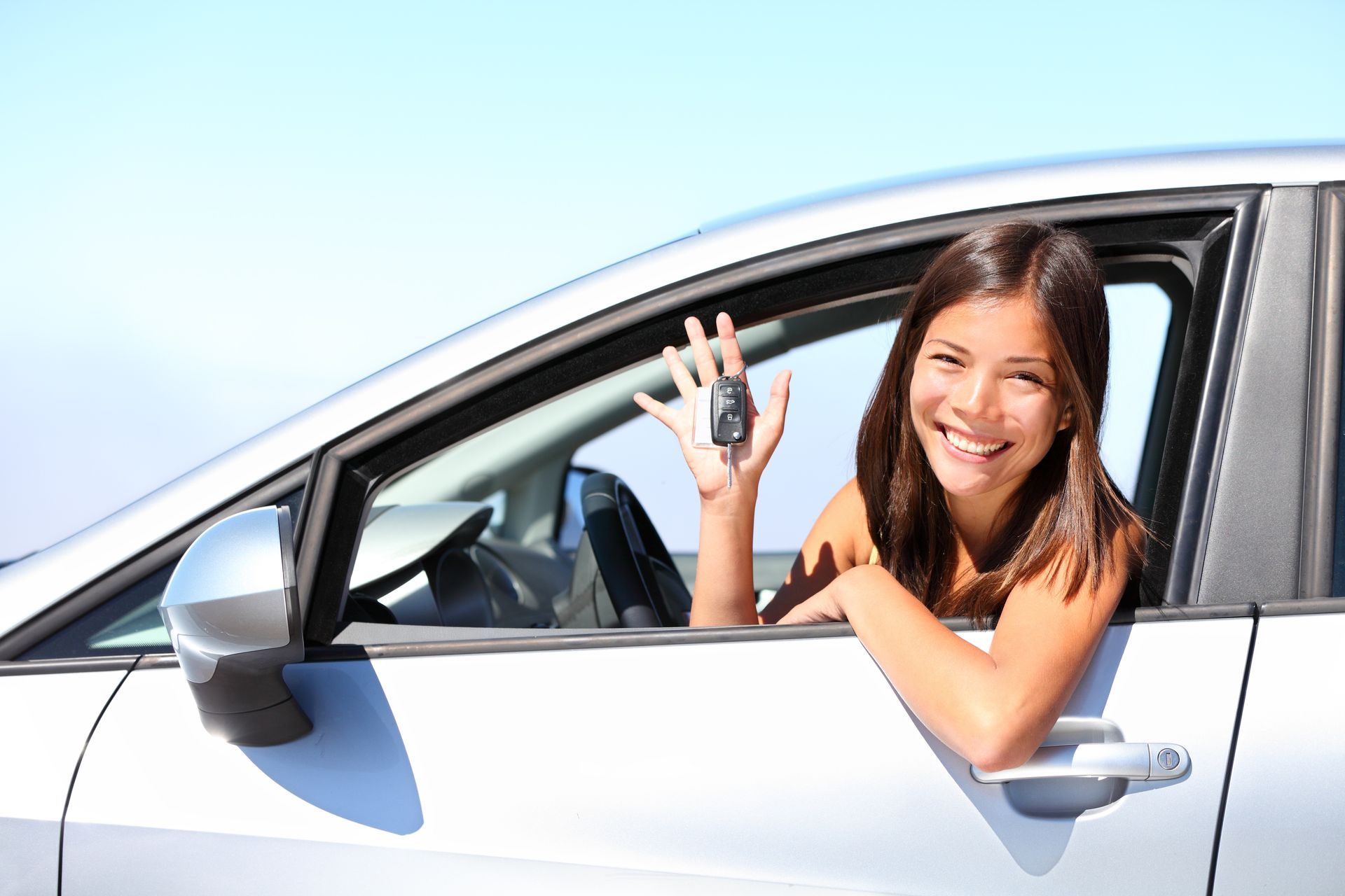 Woman in car, holding car keys, smiling. Bright blue sky background.
