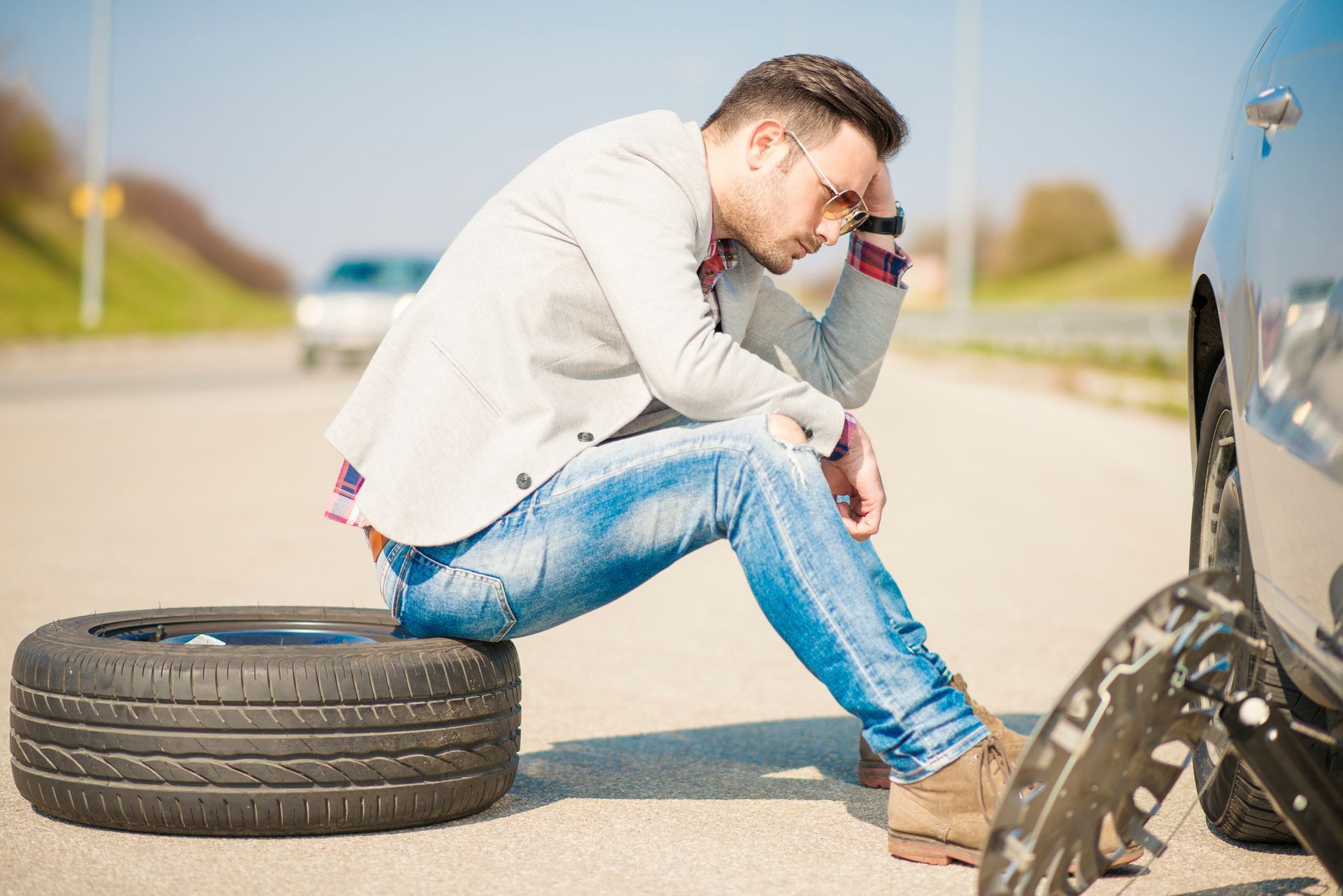 Man sitting dejectedly on a spare tire next to a car with a flat tire on a roadside.