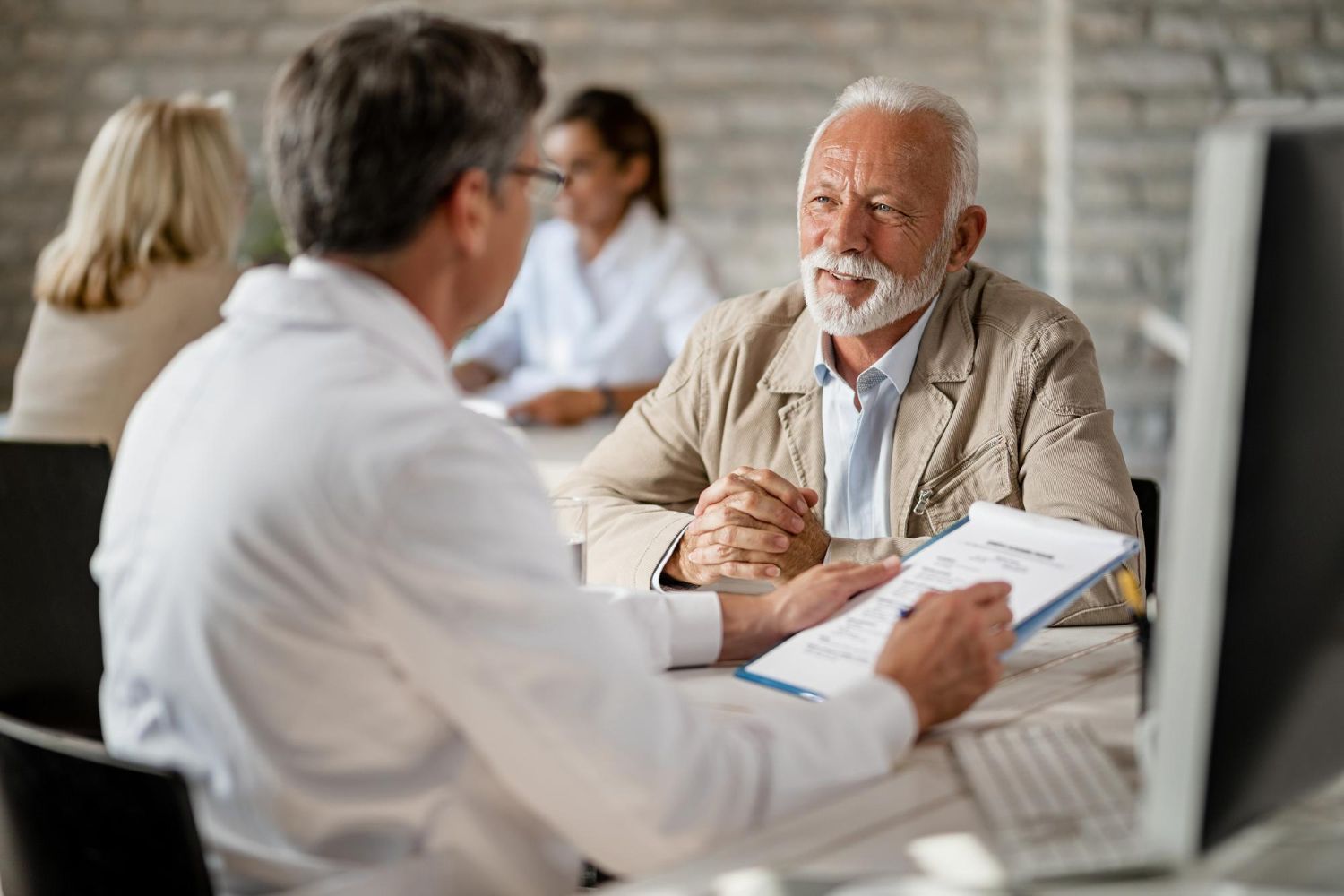 People in a bright office meeting, with one man speaking to a clinician across a table.