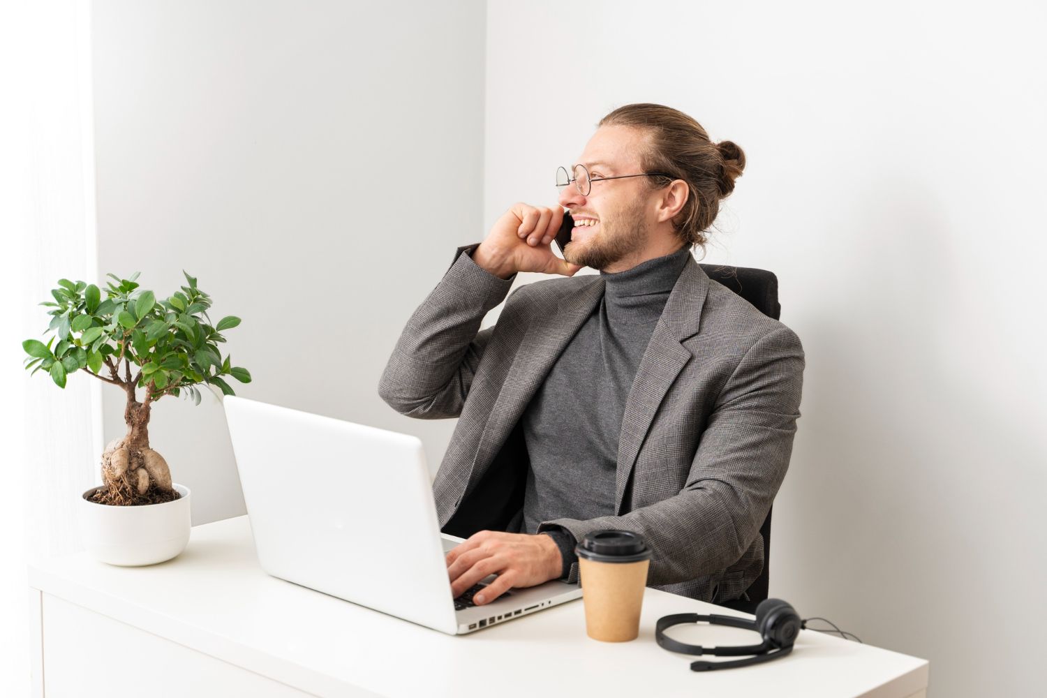 Person at a desk on the phone, using a laptop beside a coffee cup and potted plant.