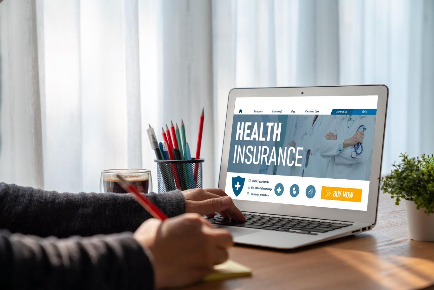 Hands writing beside a laptop showing a health insurance website on a desk