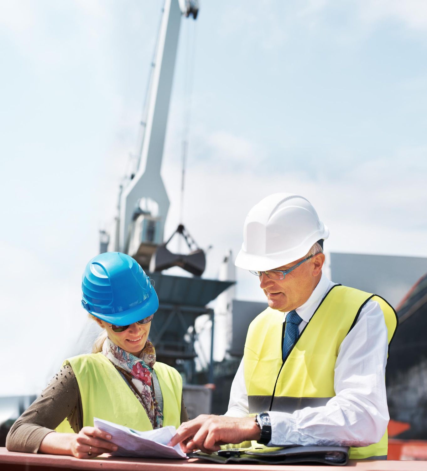 Two construction workers in hard hats reviewing plans at a job site with a crane behind them