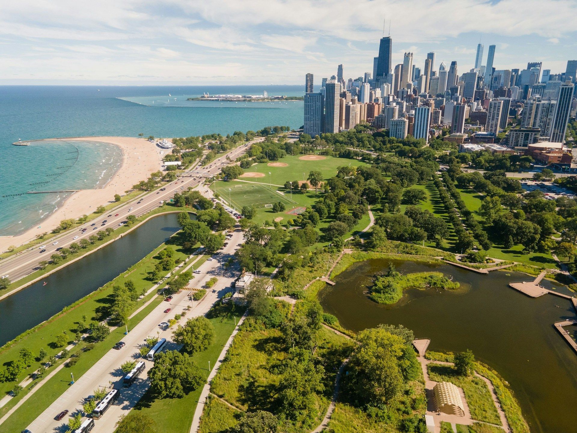 Aerial view of a sandy beach, turquoise water, green parkland, and a city skyline by the shore.