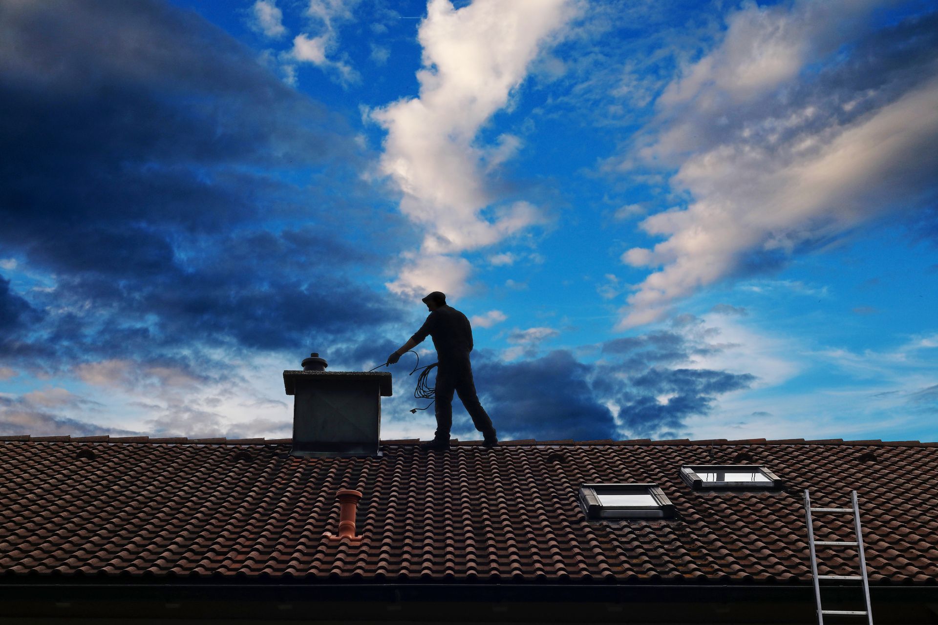 Worker standing on a tiled roof near a chimney under a dramatic blue sky.