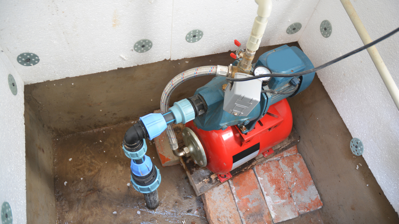 Water pump inside a concrete enclosure. The pump is red and teal, connected to black and blue pipes.