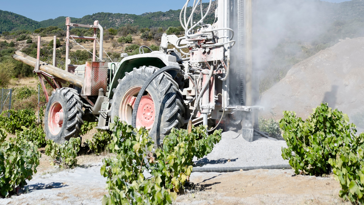 Tractor drilling a well in a vineyard, spraying water and dust, sunny day.