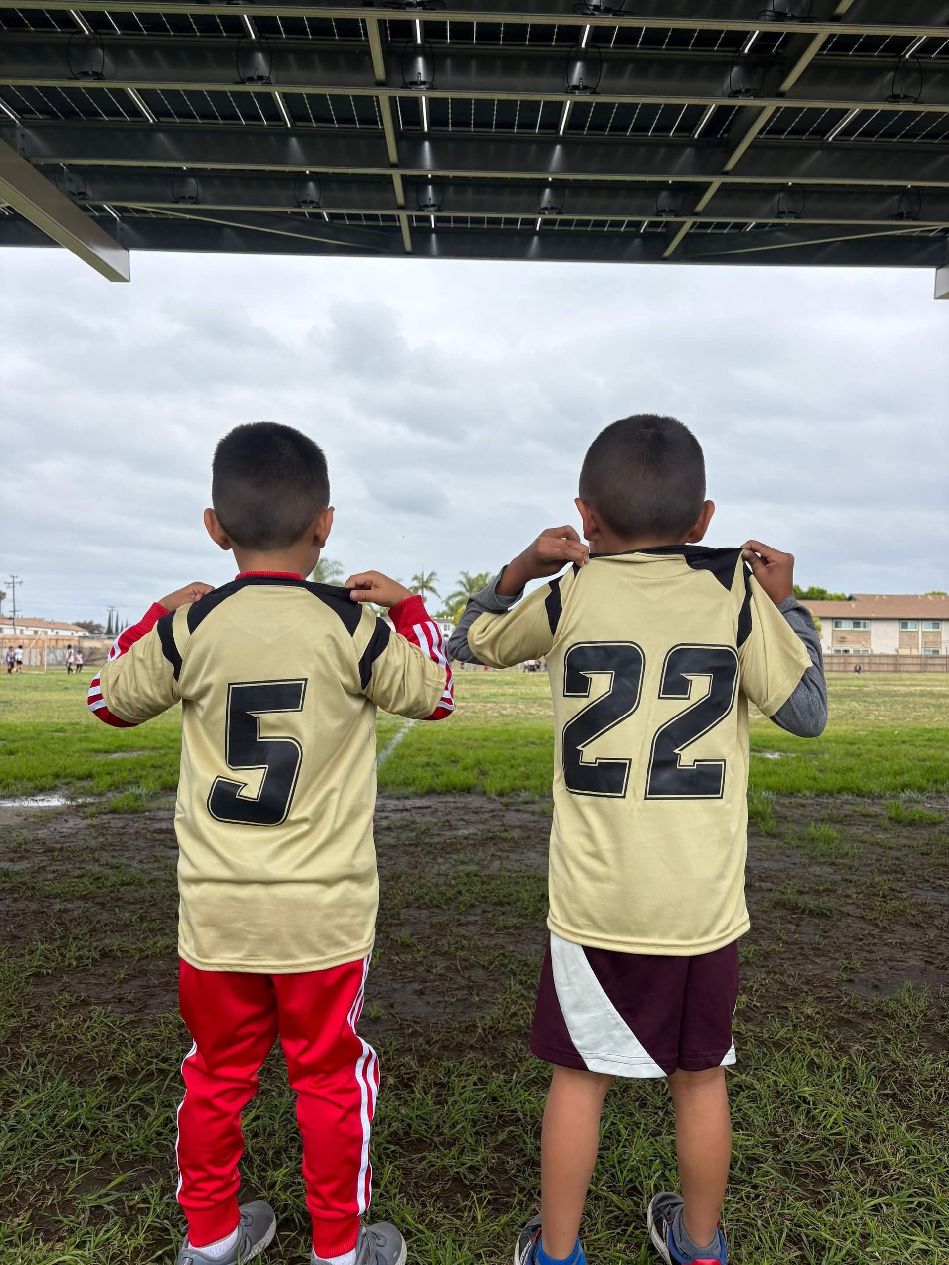 Dos niños con camisetas deportivas están de pie bajo un techo, vistos desde atrás en un campo de hierba.