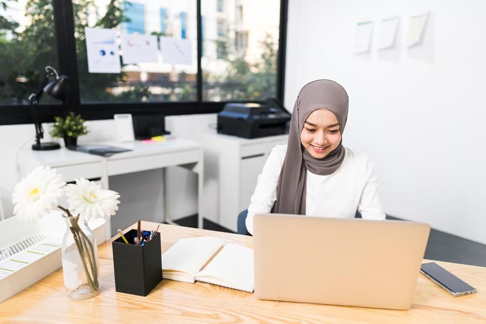 A woman in a hijab is sitting at a desk using a laptop computer.