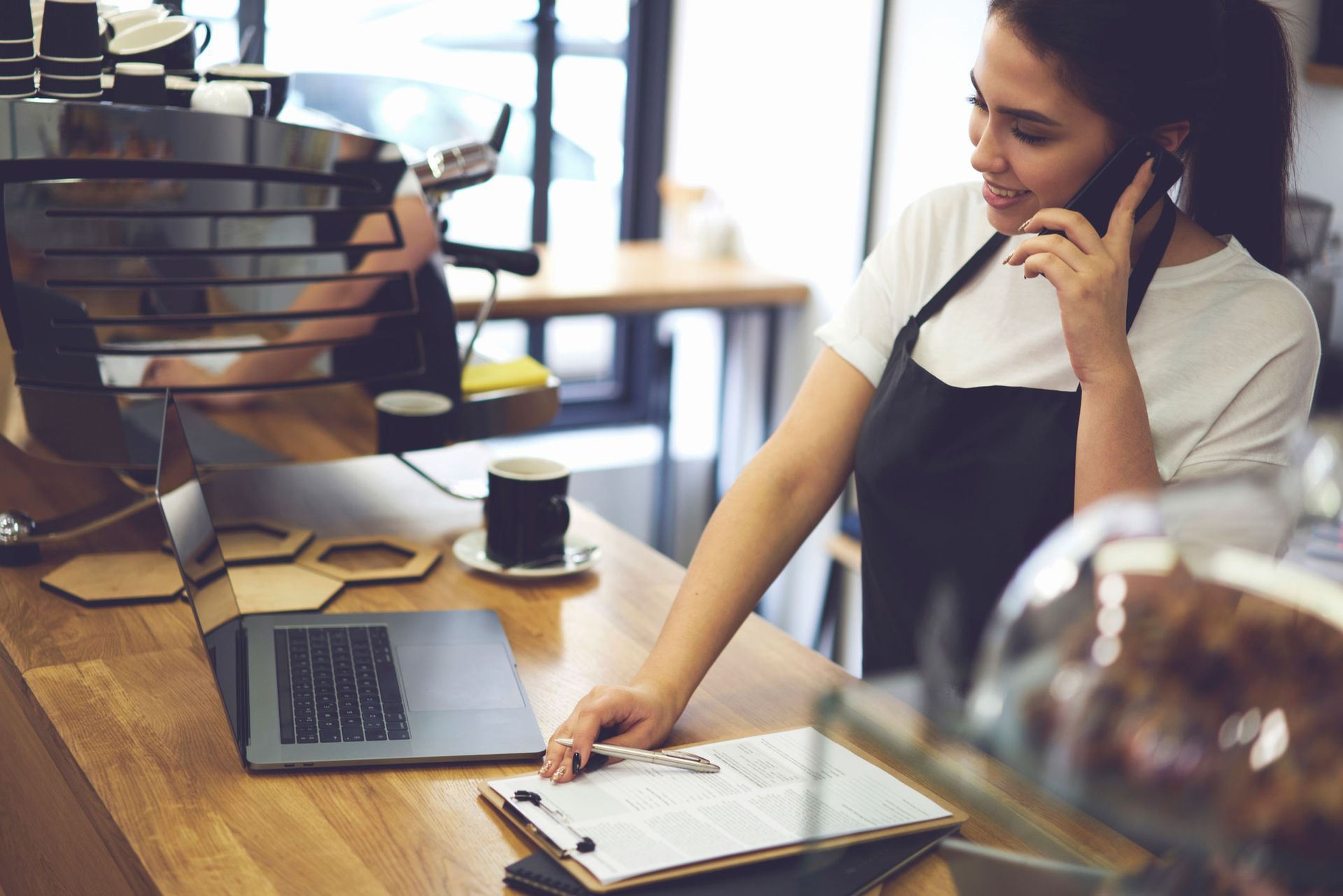 A woman in an apron is talking on a cell phone in a cafe.