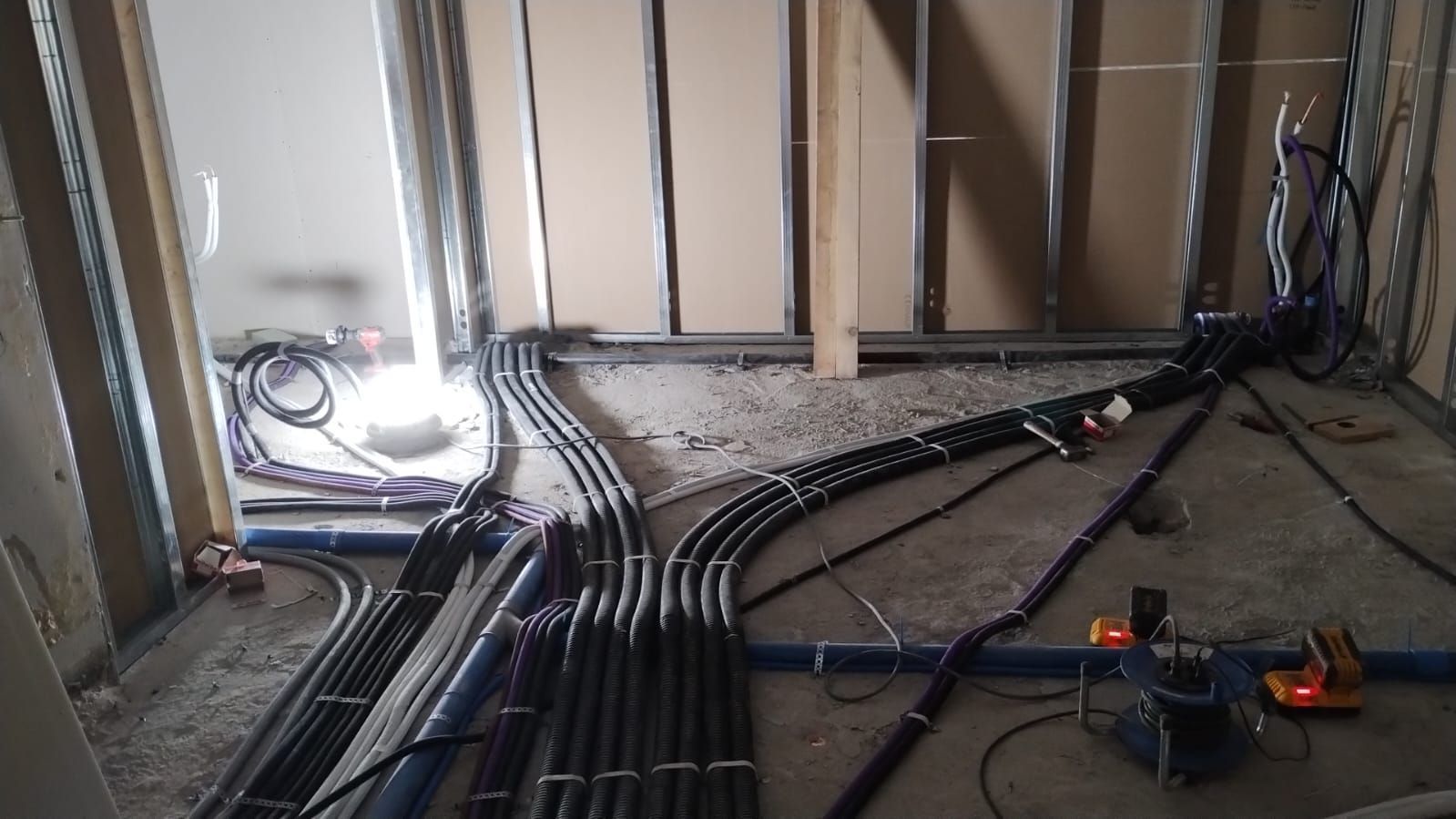 Cablaggio elettrico durante la costruzione, con condotti neri e tubi blu su un pavimento di cemento all'interno di una stanza.