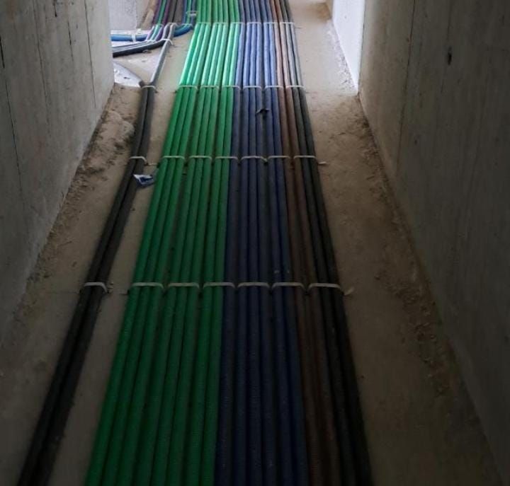 Tubi verdi, blu e marroni che corrono lungo un pavimento di cemento, in uno stretto corridoio.