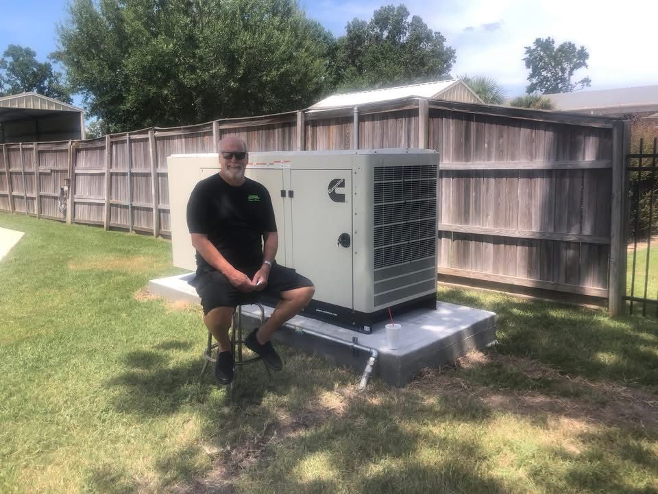 Man sits near a Cummins generator in a backyard; brown fence, green grass, sunny day.