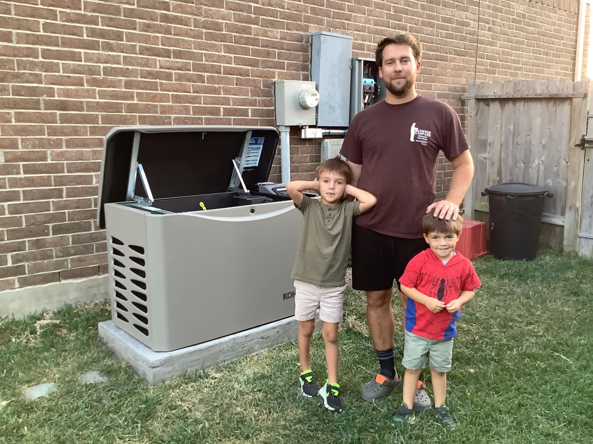 A man and two young boys pose with a standby generator by a brick wall and fence on grass.