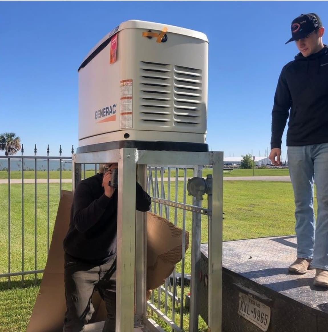 Two men installing a Generac generator outdoors. One works inside the metal cage, the other watches. Sunny day.