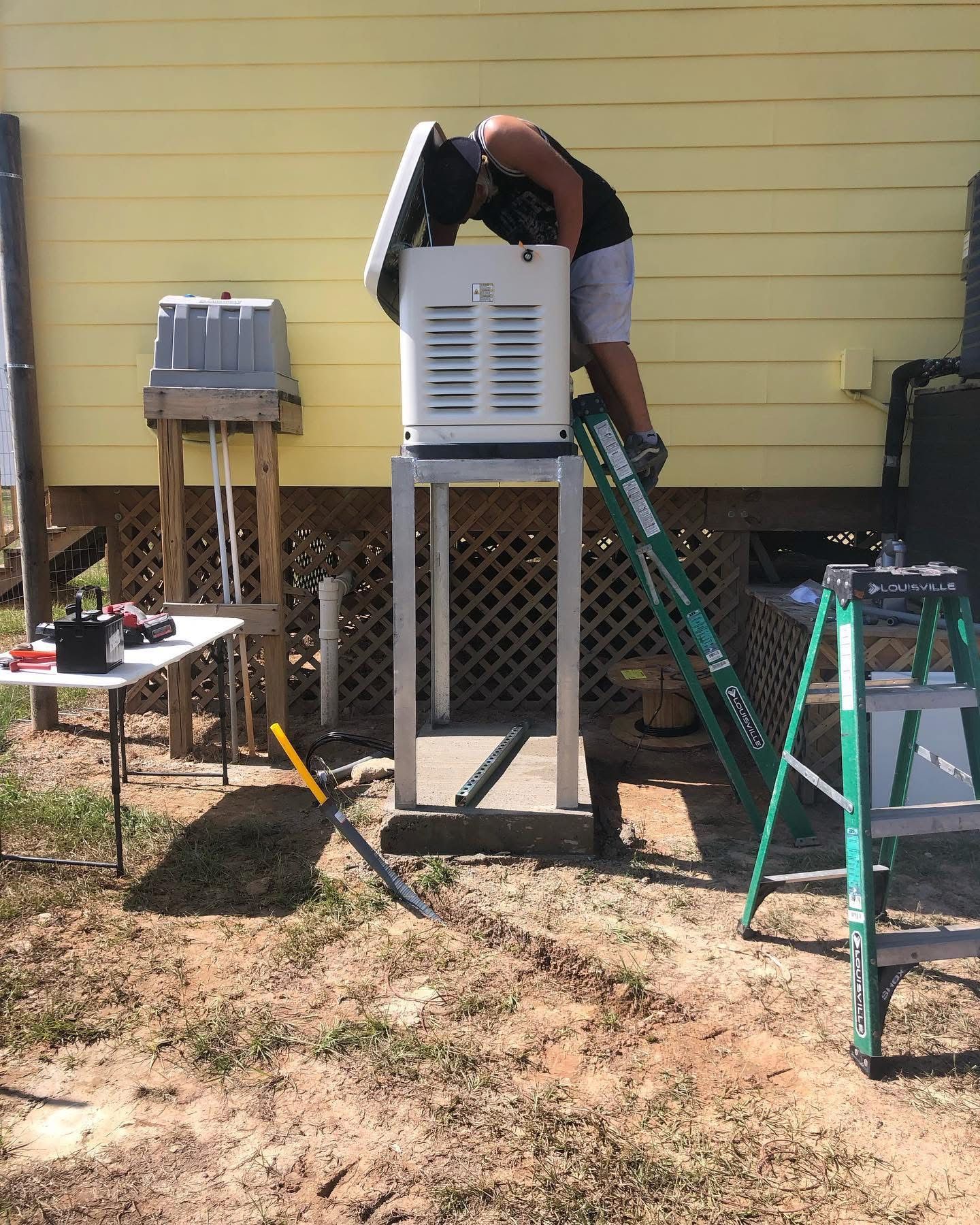 Person on ladder installing air conditioner on stand outside yellow house.