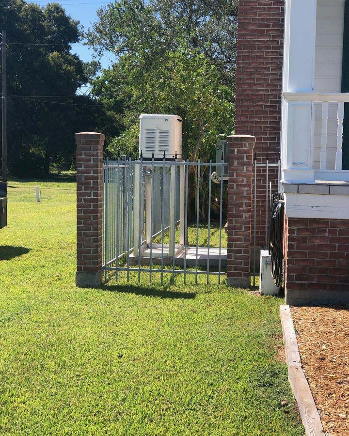 Brick pillars and metal gate enclose an electrical unit on a lawn next to a brick building.