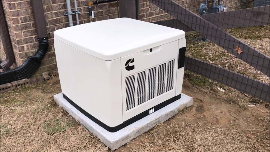 A beige Cummins home generator on a concrete pad in a yard, next to a fence and brick wall.