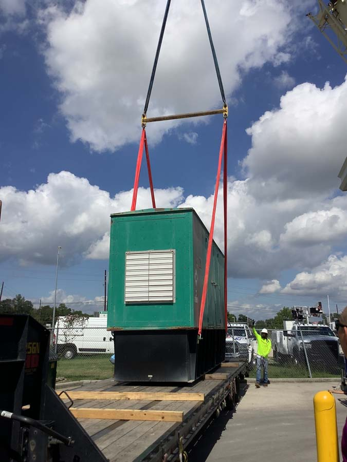 Green generator being lifted by a crane onto a flatbed truck, worker directing. Sunny day.