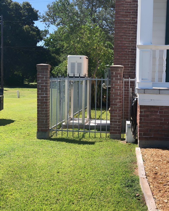 Generator behind a metal fence, brick columns, and green grass near a brick and white building.