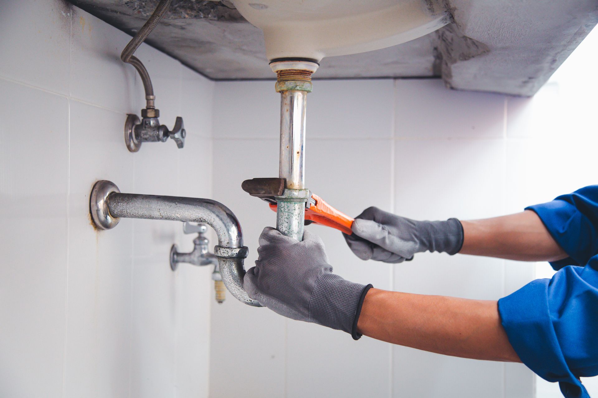 Plumber in blue work shirt and gray gloves, using a wrench on pipes under a white sink in a tiled bathroom.