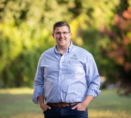 adam devitt - a man wearing glasses and a blue shirt is standing in a field with his hands in his pockets.