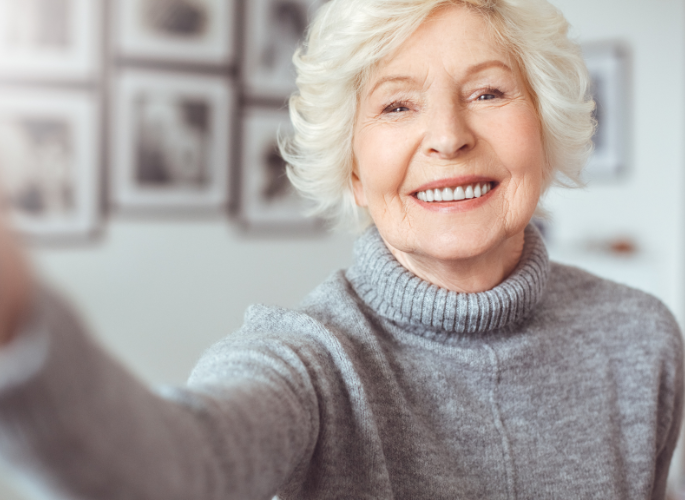 Woman smiling, taking a selfie, wearing a gray turtleneck sweater. Soft focus background of framed photos.