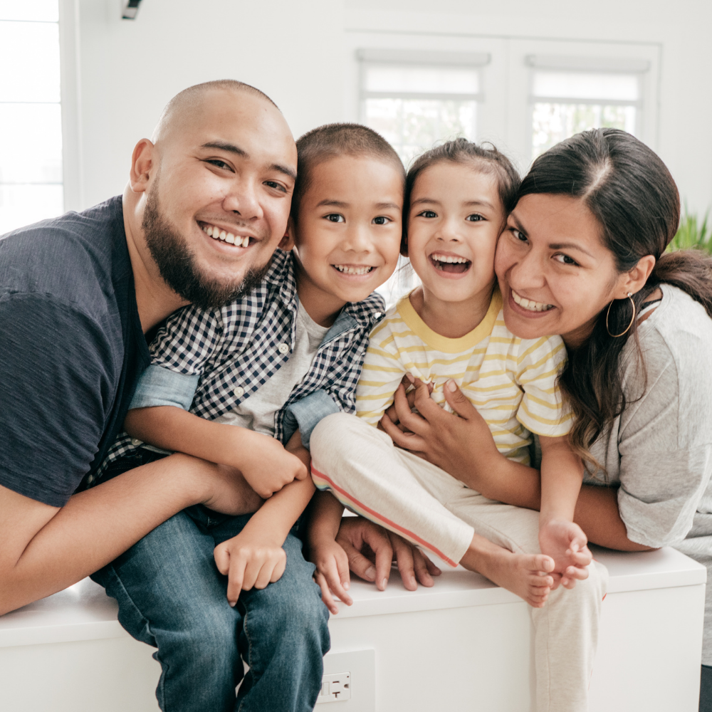 Family of four smiling, sitting close together indoors; sunny, happy expressions.
