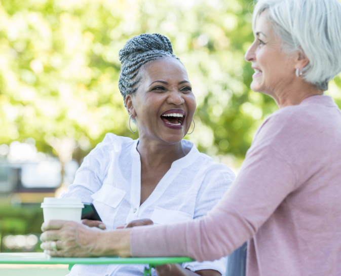 Two women laughing outdoors, one holding a coffee cup, with a blurred green background.