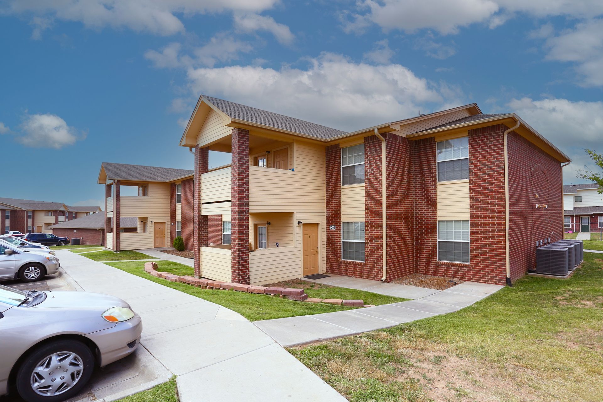 Apartment building exterior with brick and tan siding, sidewalks, and parked cars on a sunny day