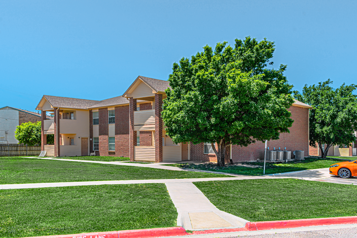 Apartment building with brick exterior, green lawn, sidewalk, and a large tree in front