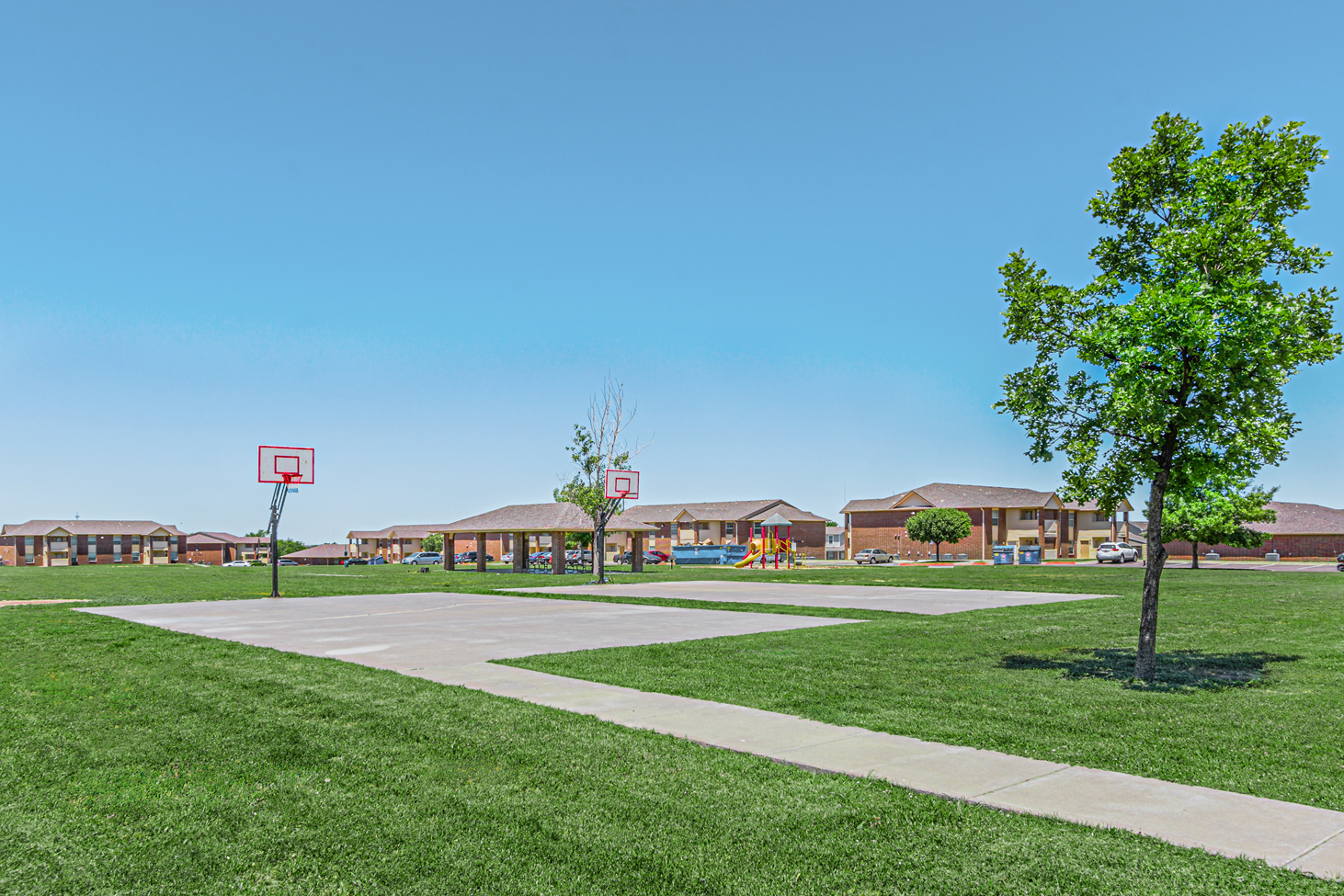 School playground with basketball hoop, sidewalk, grassy field, and suburban homes under a clear blue sky