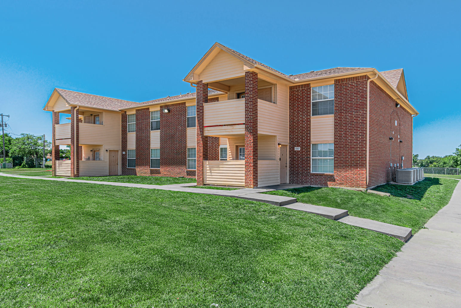 Brick apartment buildings with balconies beside a green lawn and sidewalk under a clear sky