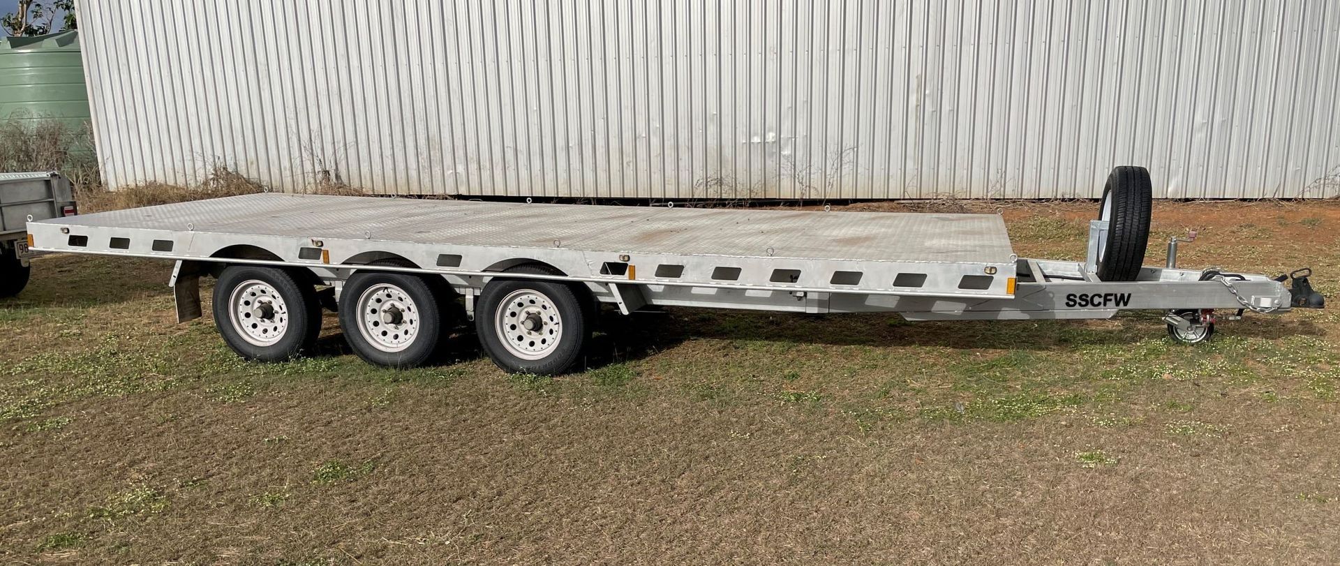 A flatbed trailer is parked in the grass in front of a white fence.