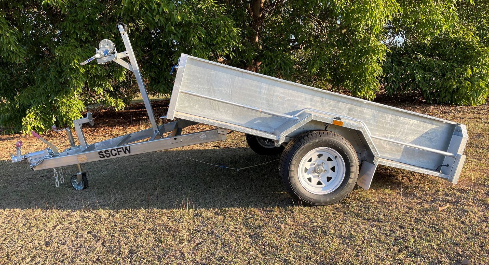 A small trailer is parked on the grass in front of trees.
