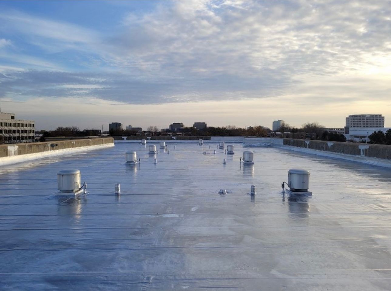 Flat commercial rooftop under a cloudy sky, featuring multiple vents and a reflective, silver coating. Distant buildings line the horizon.