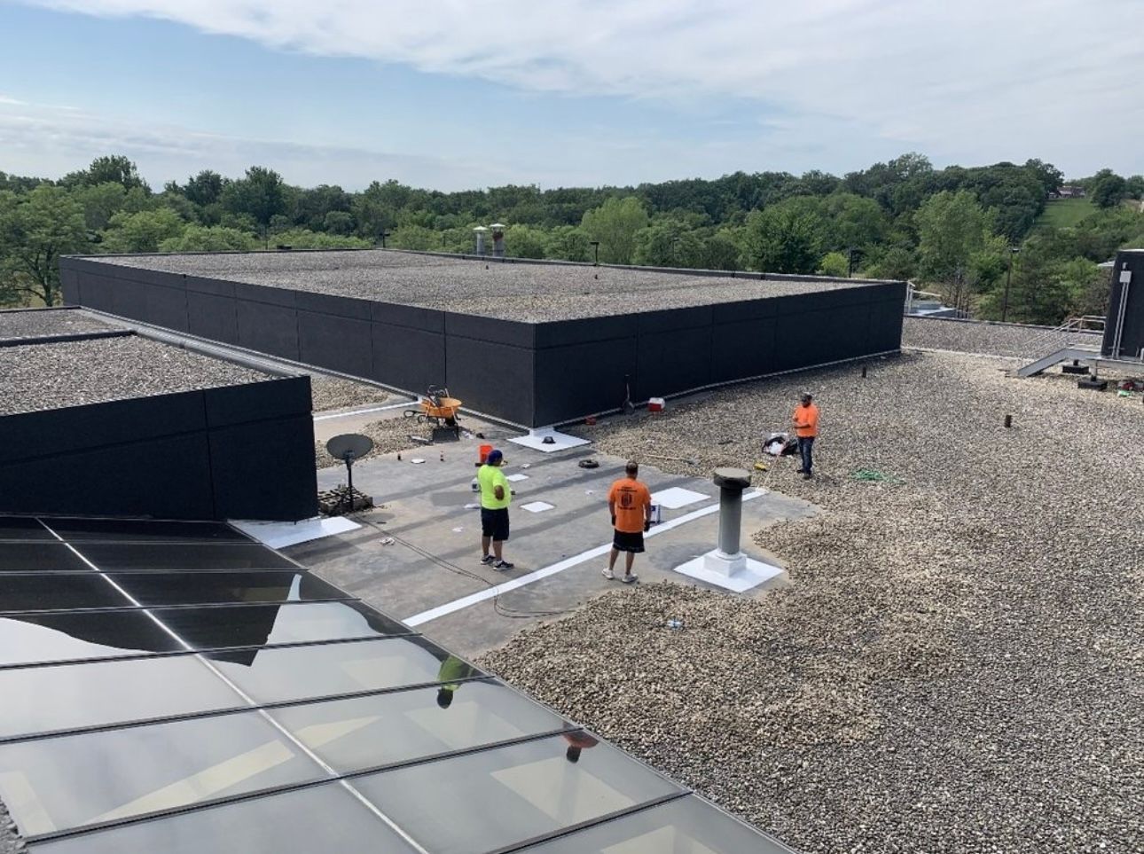 Workers on a flat rooftop, with a gravel surface, installing a black barrier, and solar panels in the foreground.
