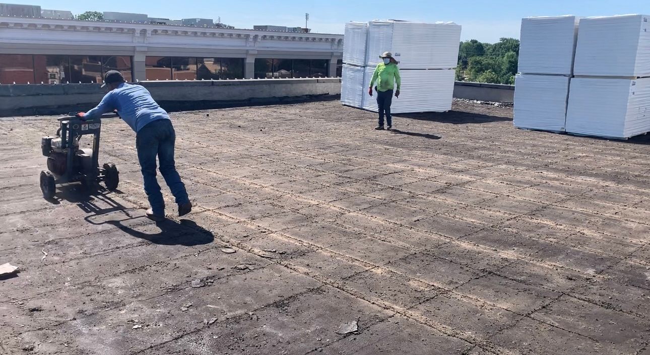Two workers on a roof, one operating a small machine, the other standing nearby near stacks of white material.